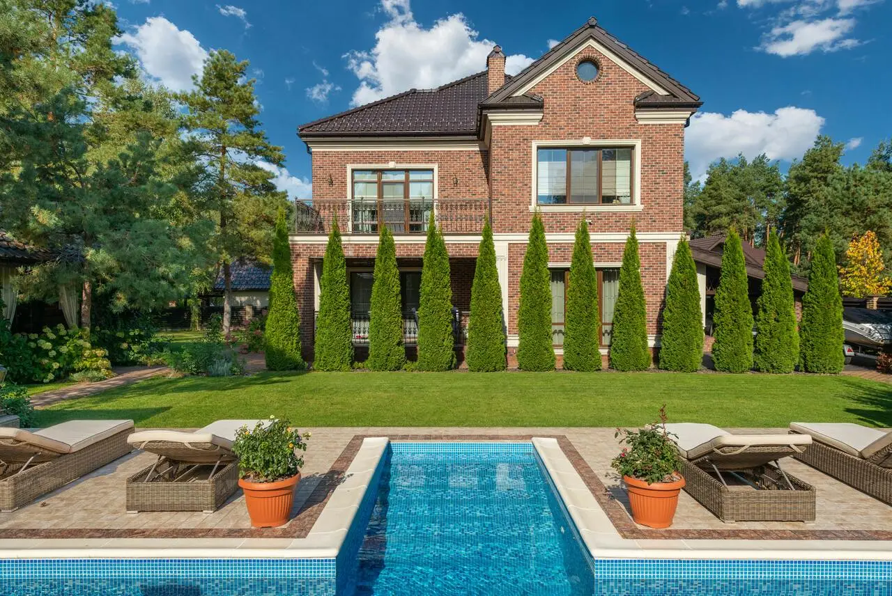 A two-story brick house with tall windows, surrounded by neatly trimmed trees and a lawn, stands behind a rectangular swimming pool with lounge chairs and potted plants on a sunny day.