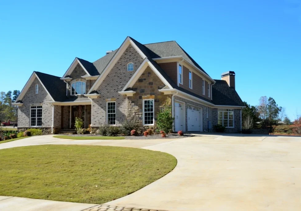 Two-story suburban house with brick and stone exterior, a three-car garage, and a manicured lawn under a clear blue sky.