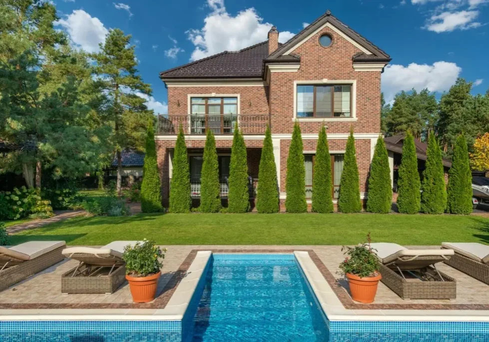 A two-story brick house with tall windows, surrounded by neatly trimmed trees and a lawn, stands behind a rectangular swimming pool with lounge chairs and potted plants on a sunny day.